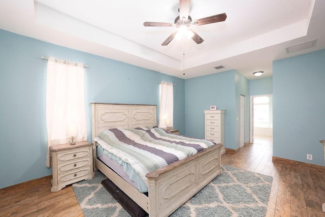 bedroom featuring light wood-type flooring, ensuite bath, a raised ceiling, and ceiling fan