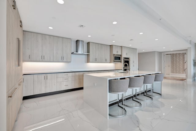 kitchen featuring light brown cabinets, an island with sink, decorative backsplash, wall chimney range hood, and stainless steel microwave