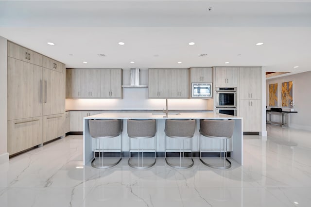 kitchen with appliances with stainless steel finishes, an island with sink, wall chimney range hood, and light brown cabinets