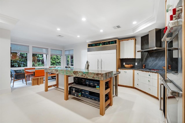 kitchen featuring appliances with stainless steel finishes, white cabinetry, decorative backsplash, a center island, and wall chimney range hood