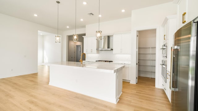 kitchen featuring appliances with stainless steel finishes, a kitchen island with sink, hanging light fixtures, and wall chimney range hood