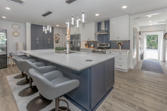 kitchen featuring stainless steel appliances, wall chimney range hood, a large island with sink, white cabinets, and light wood-type flooring