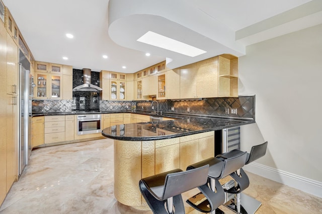 kitchen with wall chimney exhaust hood, a kitchen bar, dark stone countertops, appliances with stainless steel finishes, and kitchen peninsula