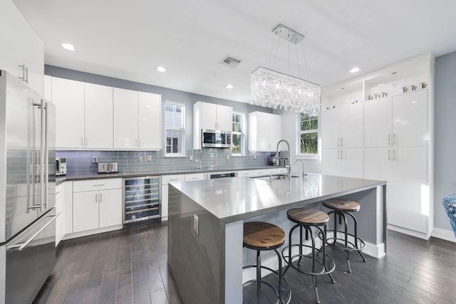 kitchen with appliances with stainless steel finishes, beverage cooler, decorative light fixtures, white cabinets, and an island with sink