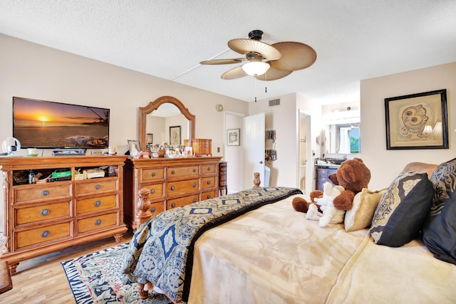 bedroom with ceiling fan, light wood-type flooring, a textured ceiling, and connected bathroom