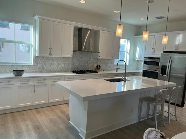 kitchen with wall chimney range hood, sink, appliances with stainless steel finishes, an island with sink, and white cabinets