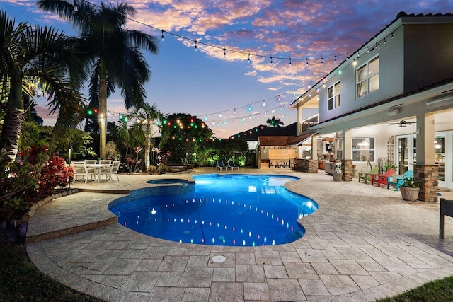 pool at dusk featuring an in ground hot tub, ceiling fan, a patio area, and exterior kitchen