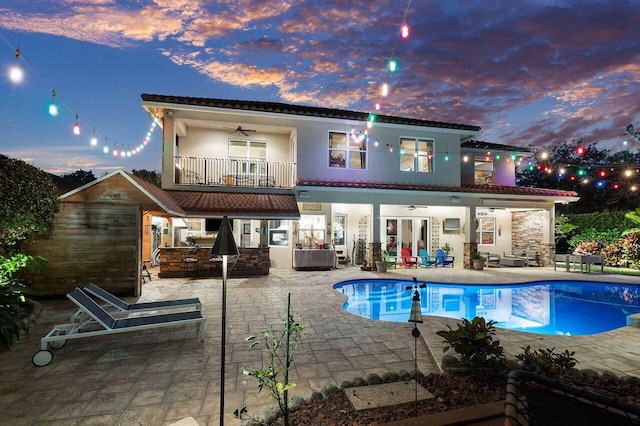 back house at dusk with ceiling fan, a balcony, an outdoor living space, and a patio