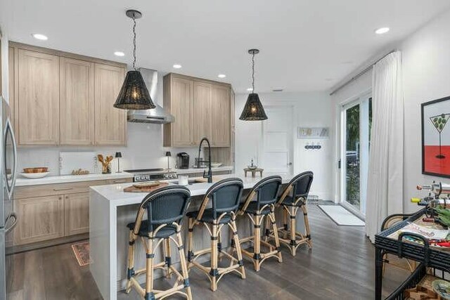 kitchen featuring light brown cabinets, wall chimney range hood, dark hardwood / wood-style floors, pendant lighting, and a kitchen island with sink