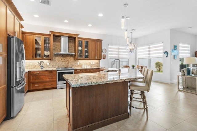 kitchen featuring pendant lighting, a kitchen island with sink, wall chimney range hood, sink, and appliances with stainless steel finishes