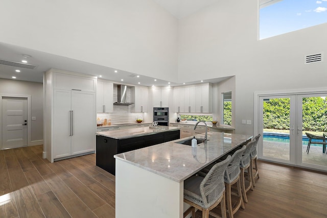 kitchen with a high ceiling, white cabinets, wall chimney exhaust hood, an island with sink, and sink