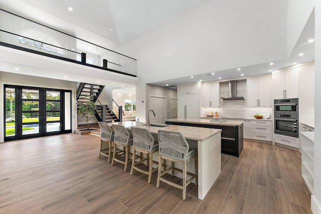 kitchen featuring light stone countertops, a high ceiling, white cabinets, wall chimney range hood, and a kitchen island with sink