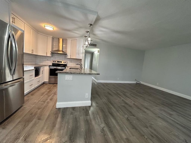 kitchen with appliances with stainless steel finishes, wall chimney range hood, a center island with sink, decorative light fixtures, and white cabinets