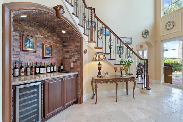 bar with light stone counters, beverage cooler, light tile patterned flooring, and high vaulted ceiling