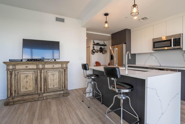 kitchen with appliances with stainless steel finishes, decorative backsplash, hanging light fixtures, a center island with sink, and light wood-type flooring