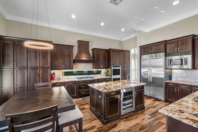 kitchen featuring a center island with sink, hanging light fixtures, built in appliances, custom range hood, and beverage cooler