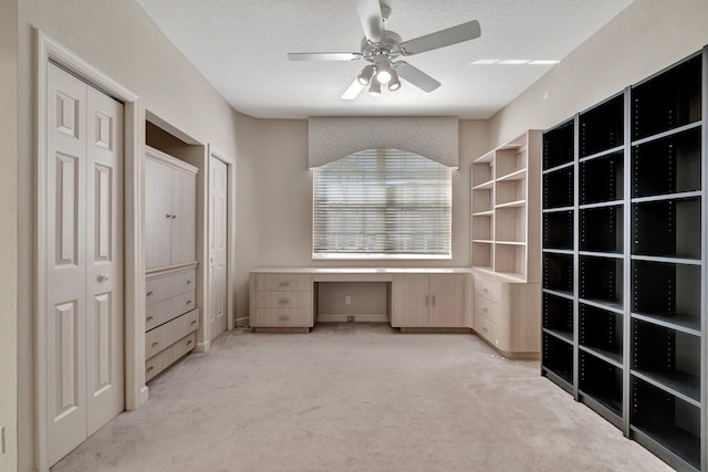 unfurnished office featuring a textured ceiling, built in desk, light colored carpet, and ceiling fan