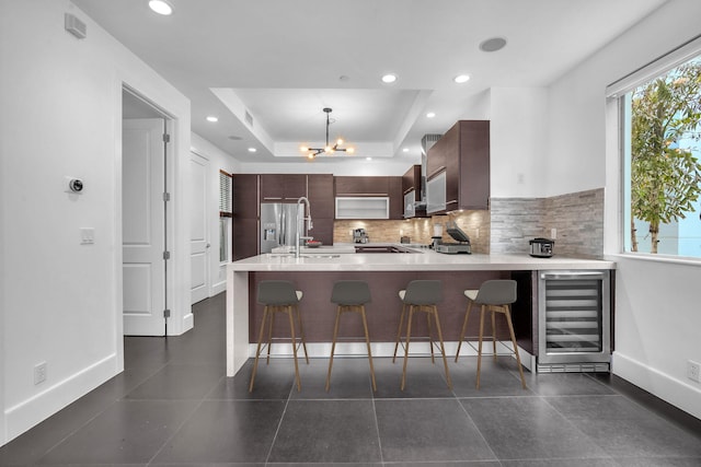 kitchen with kitchen peninsula, stainless steel fridge, a breakfast bar, a raised ceiling, and beverage cooler