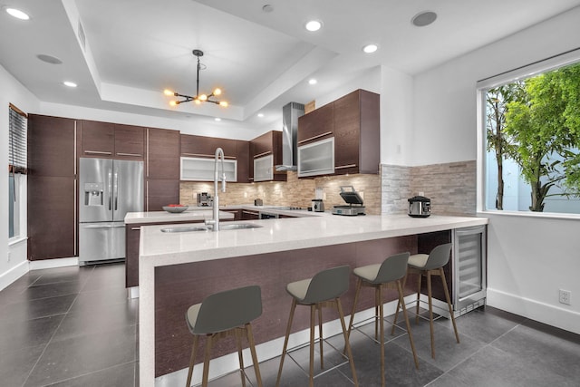 kitchen featuring kitchen peninsula, stainless steel refrigerator with ice dispenser, a breakfast bar, a tray ceiling, and hanging light fixtures