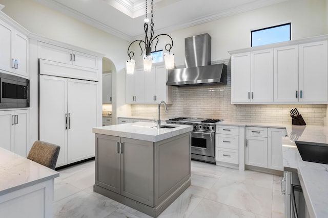 kitchen featuring built in appliances, pendant lighting, wall chimney range hood, a kitchen island with sink, and white cabinets