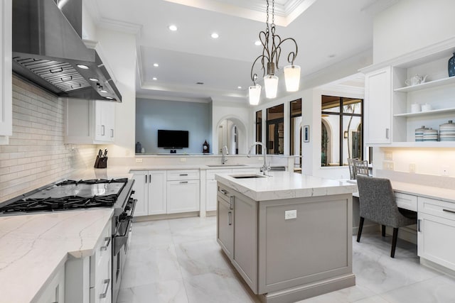 kitchen with wall chimney range hood, built in desk, an island with sink, and white cabinets