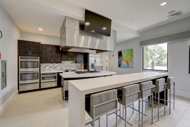 kitchen featuring a breakfast bar area, island range hood, appliances with stainless steel finishes, a kitchen island, and backsplash