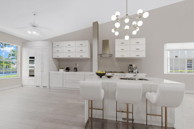 interior space with white cabinetry, wall chimney exhaust hood, hanging light fixtures, lofted ceiling, and ceiling fan with notable chandelier