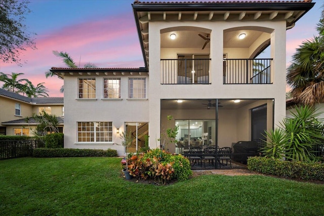 back house at dusk with a balcony, ceiling fan, a patio area, and a lawn