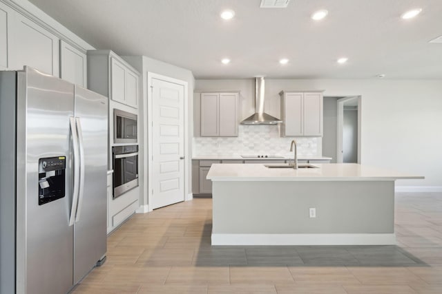 kitchen featuring gray cabinets, appliances with stainless steel finishes, an island with sink, sink, and wall chimney range hood