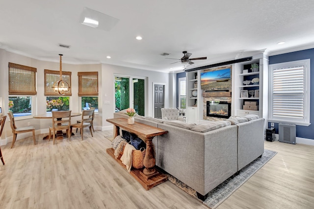 living room with built in shelves, a stone fireplace, plenty of natural light, light hardwood / wood-style floors, and ceiling fan with notable chandelier