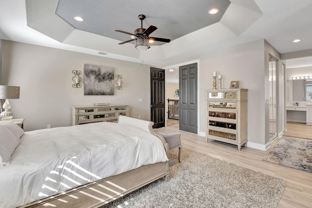 bedroom featuring a raised ceiling, ensuite bath, ceiling fan, and light hardwood / wood-style flooring