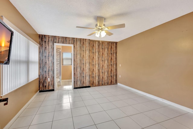 empty room with a textured ceiling, ceiling fan, light tile patterned floors, wooden walls, and baseboards