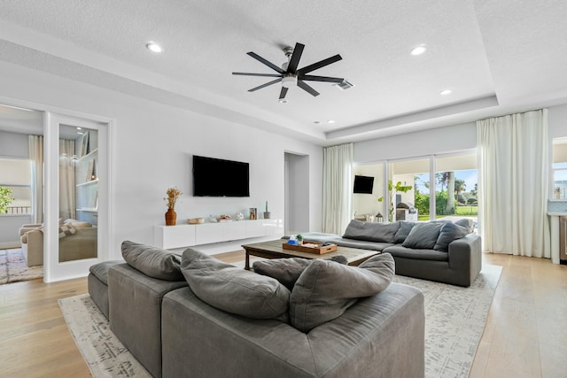 living room with a raised ceiling, a textured ceiling, light hardwood / wood-style floors, and ceiling fan with notable chandelier