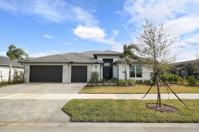 prairie-style home with decorative driveway, a tile roof, stucco siding, an attached garage, and a front lawn