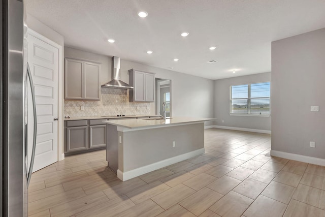 kitchen featuring decorative backsplash, stainless steel fridge, wall chimney exhaust hood, a center island with sink, and gray cabinets