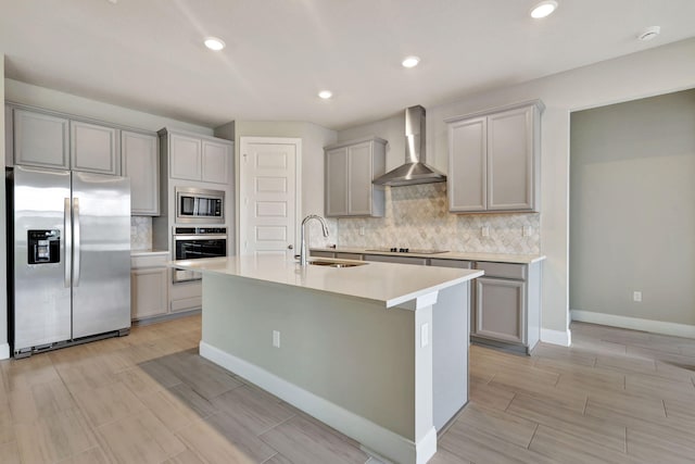 kitchen featuring gray cabinetry, wall chimney range hood, sink, an island with sink, and stainless steel appliances