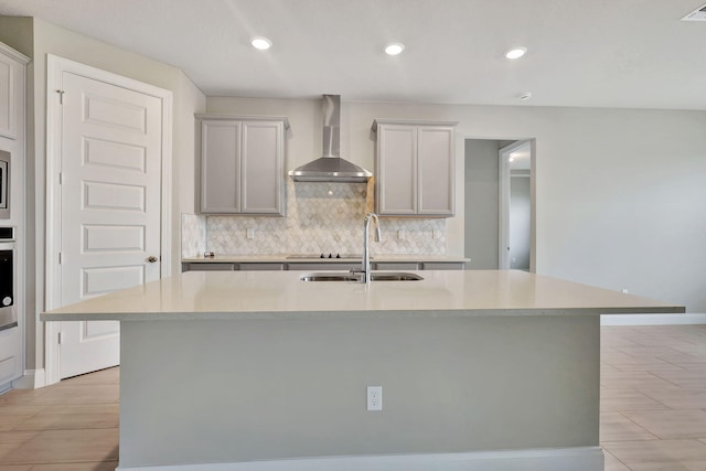 kitchen with wall chimney range hood, sink, light tile patterned floors, an island with sink, and appliances with stainless steel finishes