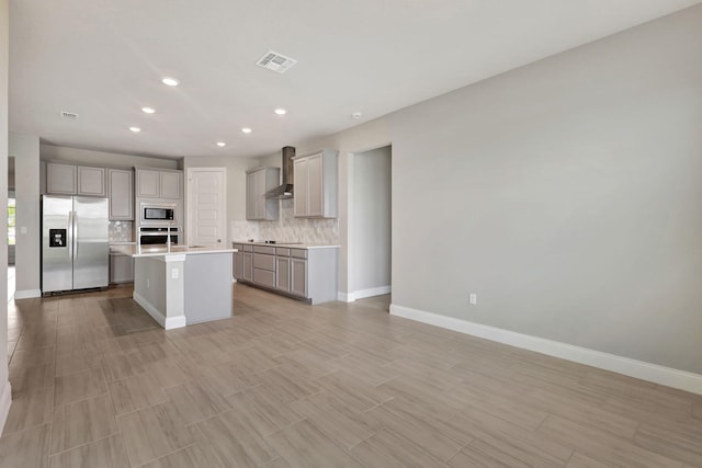 kitchen featuring a center island with sink, wall chimney range hood, gray cabinets, tasteful backsplash, and stainless steel appliances