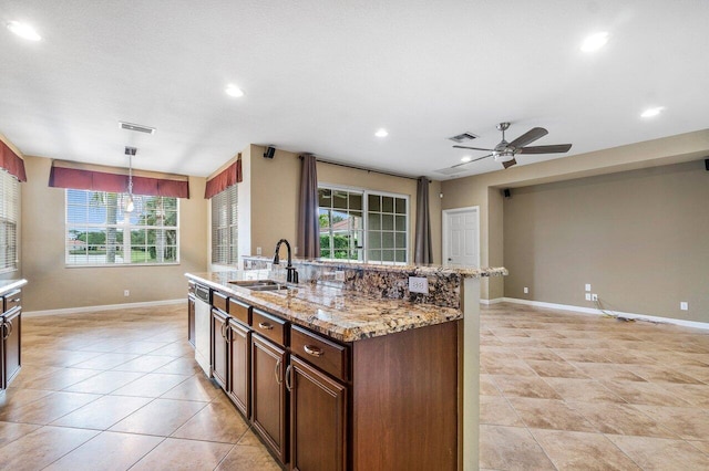 kitchen with ceiling fan, sink, light stone counters, an island with sink, and decorative light fixtures