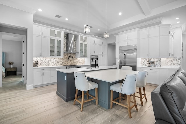 kitchen with white cabinets, built in refrigerator, a center island with sink, and wall chimney range hood