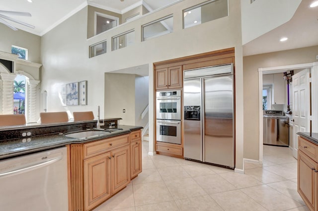 kitchen featuring stainless steel appliances, light tile patterned floors, a towering ceiling, dark stone counters, and sink