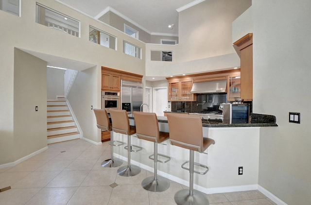 kitchen with appliances with stainless steel finishes, kitchen peninsula, light tile patterned floors, dark stone counters, and wall chimney range hood