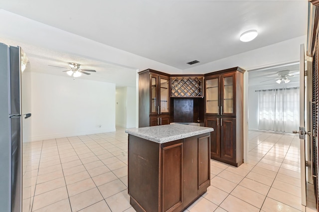 kitchen with stainless steel fridge, light stone counters, dark brown cabinetry, light tile patterned floors, and a kitchen island