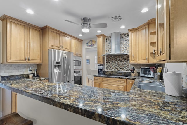 kitchen with wall chimney range hood, backsplash, kitchen peninsula, dark stone counters, and appliances with stainless steel finishes