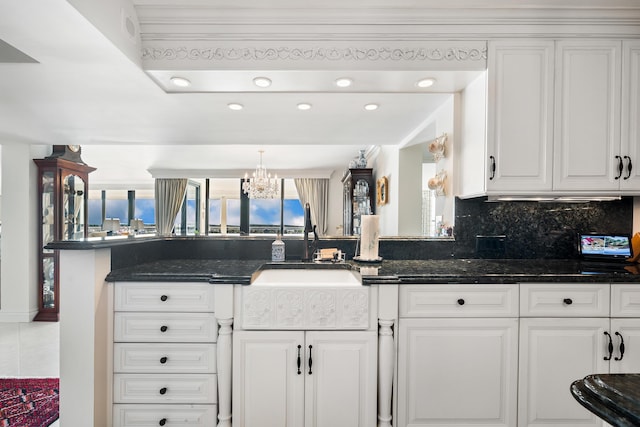 kitchen featuring an inviting chandelier, kitchen peninsula, decorative backsplash, dark stone countertops, and white cabinetry