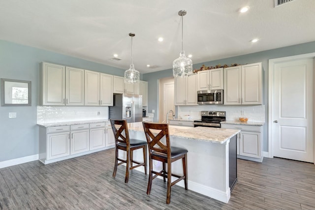 kitchen with tasteful backsplash, stainless steel appliances, decorative light fixtures, light hardwood / wood-style floors, and an island with sink