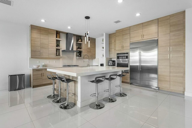 kitchen featuring a kitchen bar, wall chimney exhaust hood, stainless steel appliances, pendant lighting, and a center island with sink