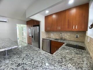 kitchen with brown cabinetry, appliances with stainless steel finishes, a wall mounted air conditioner, light stone countertops, and backsplash