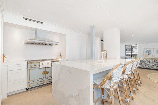 kitchen featuring white cabinets, light stone counters, high quality stove, a kitchen island with sink, and wall chimney range hood