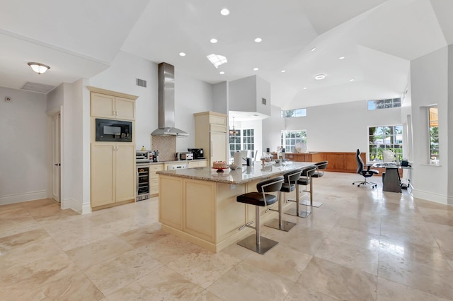 kitchen with wall chimney range hood, a kitchen bar, a towering ceiling, an island with sink, and black microwave